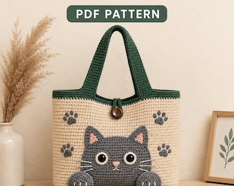 May include: A crochet tote bag featuring a beige body with a dark green top border and handles. The bag displays a dark gray cat face with pink inner ears and white whiskers, accented by four dark gray paw prints and a single brown button closure.