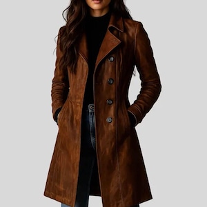 May include: A long, brown leather coat featuring a notched lapel, long sleeves, and a front button closure with four visible dark buttons. The coat has a tailored fit with vertical seam detailing and two side pockets, worn over a black top and blue jeans.