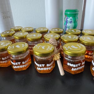 Könnte beinhalten: Kleine sechseckige Gläser gefüllt mit goldenem Honig, mit goldenen Deckeln, die mit den Buchstaben MH und einem Babyfußabdruck bedruckt sind. Die Gläser haben weiße Etiketten mit der Aufschrift SWEET BABY JAKUPKO. Ein Honiglöffel mit Goldbiene.