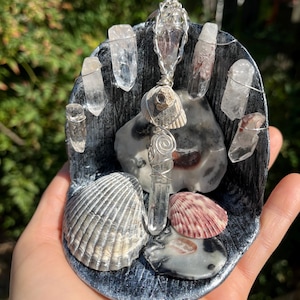 May include: A handcrafted, dark gray, shell-shaped display featuring clear quartz crystals, seashells, and a wire-wrapped crystal pendant. The display includes a variety of seashells in shades of white, gray, and pink.