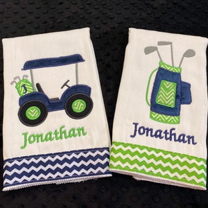 May include: Two white burp cloths with green and blue chevron trim. One cloth features an embroidered golf cart with a green golf bag and clubs. The other cloth features an embroidered golf bag with clubs. Both cloths have the name "Jonathan" embroidered in green.