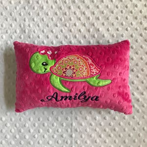 May include: A bright pink rectangular pillow with a green turtle design. The turtle has a patterned shell and a pink bow. The name "Amilya" is embroidered in black script below the turtle. The pillow is made of a soft, textured fabric.