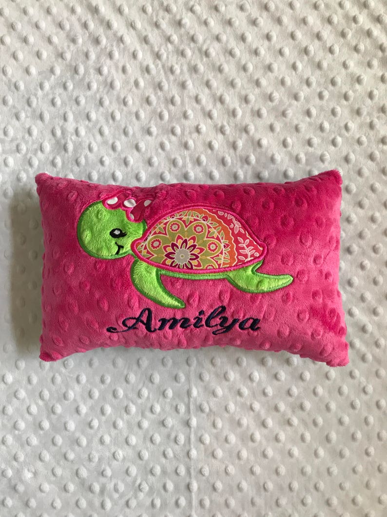 May include: A bright pink rectangular pillow with a green turtle design. The turtle has a patterned shell and a pink bow. The name "Amilya" is embroidered in black script below the turtle. The pillow is made of a soft, textured fabric.