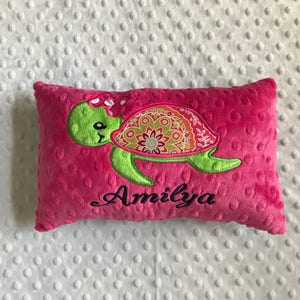 May include: A pink, rectangular pillow with a green turtle design. The turtle has a patterned shell with orange, green, and yellow accents. The name "Amilya" is embroidered in black script below the turtle.
