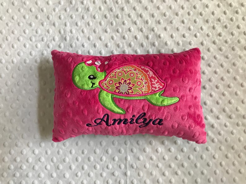 May include: A pink, rectangular pillow with a green turtle design. The turtle has a patterned shell with orange, green, and yellow accents. The name "Amilya" is embroidered in black script below the turtle.