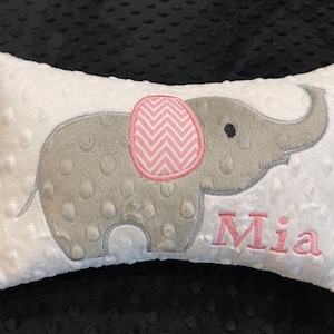 May include: A white plush pillow with a gray embroidered elephant with pink ears and a pink and white chevron pattern on the ear. The name Mia is embroidered in pink below the elephant.