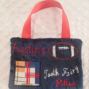 May include: A blue plush tooth fairy pillow with a red handle. The pillow has a football, plaid fabric, and the text "Austin's Tooth Fairy Pillow".