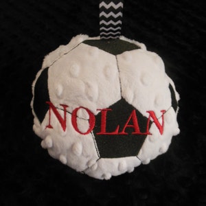May include: White and black soccer ball plush toy with the name "NOLAN" embroidered in red. The toy has a black and white chevron ribbon attached.