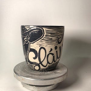 May include: A ceramic mug with a cream-coloured base and black hand-carved linocut-style illustrations. The design features a stylised cow head and the text "Sláinte" written in a bold, flowing script. The mug has a tapered shape and a smooth finish.