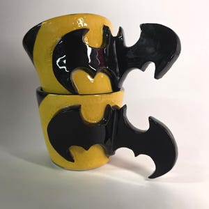 May include: Two stacked ceramic mugs with a glossy yellow finish and a prominent black handle shaped like a bat silhouette. The mugs feature a smooth, reflective surface and a distinct, stylised bat-wing design that serves as the functional handle.