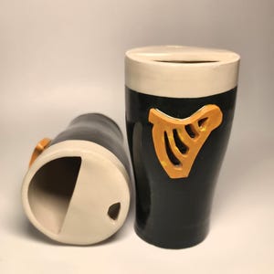 May include: Two ceramic tumblers with a black body and a white rim. Each tumbler features a raised, orange, harp-shaped emblem on the front. The design mimics a pint glass, with a wide opening and a smooth, glossy finish on the exterior surface.