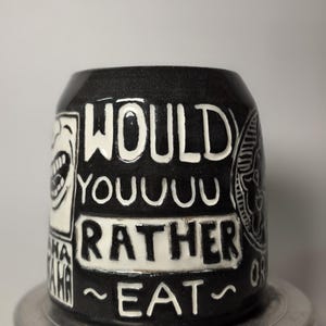 May include: A black ceramic mug with white carved lettering that reads WOULD YOUUUU RATHER EAT. The mug features additional white graphic illustrations of a laughing mouth and a circular emblem on the sides, set against a dark background.