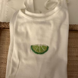 May include: A white sleeveless tank top featuring a small, centered lime wedge graphic. The lime wedge is crafted with green and light yellow beads, creating a textured, shimmering appearance against the plain white fabric of the garment.