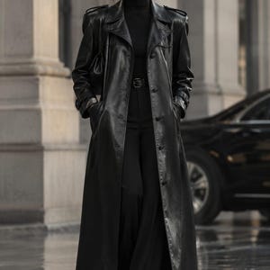 May include: A long black leather trench coat with a wide collar, shoulder epaulets, and button closures, worn over a black turtleneck sweater and black trousers with a belt. The outfit includes dark sunglasses and a small black handbag.