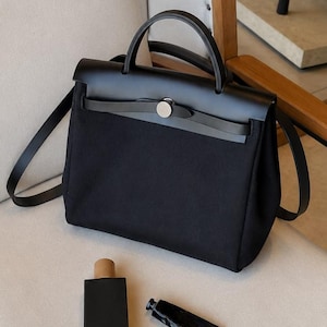 Peut inclure: Un sac à main noir doté d'une poignée supérieure structurée et d'un rabat fermé par un fermoir circulaire argenté. Le sac présente un panneau supérieur en cuir lisse et un corps en tissu texturé. À côté se trouvent un flacon noir et un tube.