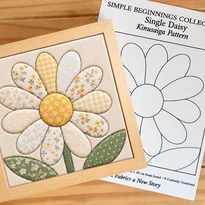 May include: A framed Kinusaiga fabric art piece featuring a daisy with yellow, white, and green patterned petals and a central yellow circle. Beside it is a paper pattern labeled SIMPLE BEGINNINGS COLLECTION Single Daisy Kinusaiga Pattern.