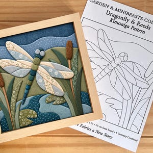 Puede incluir: Una pieza de arte en tela enmarcada con una libélula y juncos, acompañada de una hoja de patrón impresa titulada GARDEN & MINIBEASTS COLLECTION Dragonfly & Reeds Kinusaiga Pattern, que especifica un tablero de espuma de 20 x 20 cm.