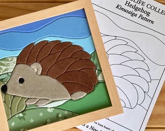 Puede incluir: Un marco cuadrado de madera clara que contiene una manualidad de tela de erizo con secciones texturizadas en marrón, beige y verde sobre un cielo azul. Al lado hay una hoja de patrón con el título BRITISH WILDLIFE COLLECTION Hedgehog Kinusaiga Pattern.