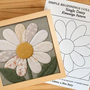 May include: A framed Kinusaiga fabric art piece featuring a daisy with white, cream, and floral patterned petals and a textured yellow center, accompanied by a paper pattern sheet titled Simple Beginnings Collection Single Daisy Kinusaiga Pattern.