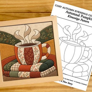 Puede incluir: Una pieza de arte en tela Kinusaiga enmarcada que representa una taza humeante sobre una pila de mantas, acompañada de una hoja de patrón titulada Cosy Autumn Evenings Collection Autumnal Pumpkin. El diseño presenta tonos tierra.