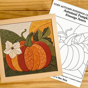 Peut inclure: Une œuvre d'art en tissu encadrée de forme carrée représentant une citrouille avec des segments à motifs floraux orange, rouges et jaunes, une fleur blanche et des feuilles vertes. À côté se trouve un patron papier intitulé COSY AUTUMN EVENINGS COLLECTION.