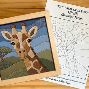 Puede incluir: Una pieza de arte en tela Kinusaiga enmarcada que representa una jirafa en un paisaje con cielo azul y colinas verdes, acompañada de una hoja de patrón impresa titulada THE WILD COLLECTION Giraffe Kinusaiga Pattern para tablero de espuma de 20 x 20 cm.