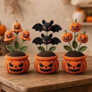 May include: Three crocheted Halloween decorations in orange pumpkin-shaped pots with jack-o'-lantern faces. The left pot holds orange bell-shaped flowers, the center pot features two black bats, and the right pot contains three round orange pumpkins.