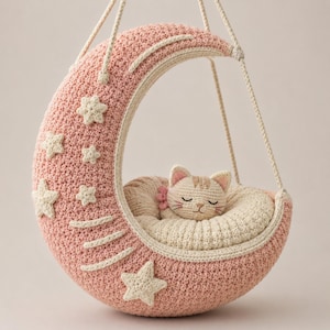 May include: A crescent moon-shaped crochet swing in dusty pink with cream-colored stars and a cream-colored inner cushion. A small, cream-colored crocheted cat with closed eyes and pink ear details rests inside the moon, suspended by cream ropes.