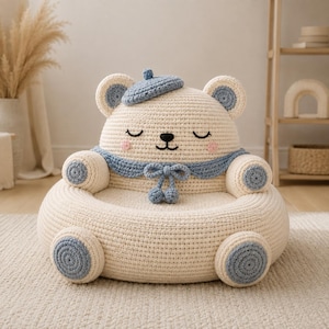 Pode incluir: Uma cadeira de crochê cor de creme em forma de urso, com uma boina azul clara, um colarinho de marinheiro a condizer com um laço e bochechas bordadas em rosa. O urso tem olhos fechados, um nariz preto e detalhes circulares azul claros nas patas.