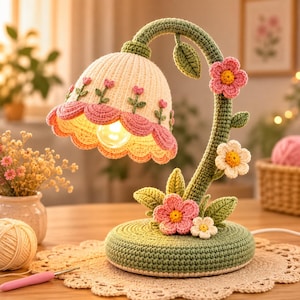 May include: A table lamp featuring a crocheted cream-colored shade with pink scalloped edges and small embroidered flowers. The base and curved stem are covered in green crochet, decorated with crocheted pink and white flowers and leaves.