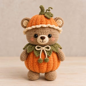 Könnte beinhalten: Ein gehäkelter Teddybär in einem Kürbis-Outfit. Der Bär besteht aus hellbraunem Garn, hat schwarze Augen und rosa Wangen. Er trägt eine orangefarbene Kürbismütze mit grünem Stiel und Blatt sowie einen orangefarbenen Kürbiskörper mit grünem Blätterkragen.