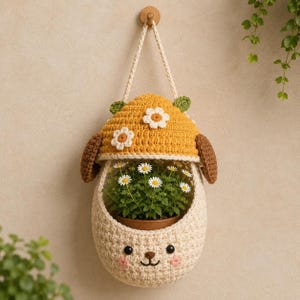 Puede incluir: Una maceta colgante de ganchillo con forma de oso que lleva un sombrero amarillo con margaritas blancas aplicadas. El oso tiene orejas marrones, ojos de botón negros, nariz bordada y mejillas rosadas. Una pequeña planta con flores blancas está dentro.