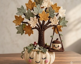 May include: A crochet sculpture featuring a cream-coloured pumpkin base with a smiling face, supporting a brown tree trunk with multi-coloured maple leaves in shades of green, yellow, and cream. A small acorn character sits on a hanging swing.