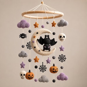 Puede incluir: Un móvil colgante con elementos de Halloween tejidos a ganchillo suspendidos de un marco de madera circular. El diseño incluye una luna creciente color crema con un murciélago negro, rodeado de calabazas naranjas, calaveras blancas, arañas negras, telarañas, nubes moradas y estrellas.