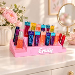 Könnte beinhalten: Ein rosafarbener, abgestufter Präsentationsständer mit dem Namen Emily in weißer Schreibschrift, der mehrere Essence Juicy Bomb Lipgloss-Tuben in verschiedenen Farben wie Rot, Orange, Blau und Lila in Reihen auf einer Kommode präsentiert.