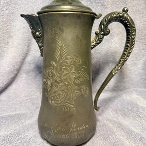 May include: A tarnished silver-toned metal coffee pot with a lid, featuring an ornate handle and a spout with decorative detailing. The body is engraved with a floral pattern and the inscription "Mr & Mrs Jordan X-mas 1900".