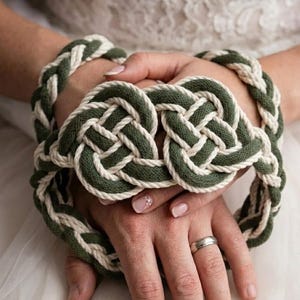 Könnte beinhalten: Ein Handfasting-Seil mit einem geflochtenen Design aus dickem olivgrünem und cremefarbenem Seil. Das Seil ist in der Mitte zu zwei markanten, symmetrischen Knoten im keltischen Stil gebunden, wobei das geflochtene Seil eine kreisförmige Schlaufe bildet.