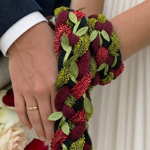 May include: A decorative braided cord featuring thick strands in deep burgundy, black, and olive green. The braid is embellished with small, light green fabric leaves tucked between the woven sections, creating a textured, multi-colored rope design.