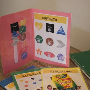 May include: A pink educational folder titled SHAPE MATCH with various colored shapes and labels. Below are additional file folder game sheets, one featuring a box of Crayola crayons, and a green spiral-bound notebook. The materials are displayed together.