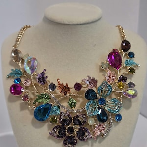 May include: A gold-tone statement necklace featuring an intricate botanical design with multi-colored faceted crystals and rhinestones. The piece includes floral and leaf motifs in shades of purple, blue, pink, green, and clear, set on a metallic frame.