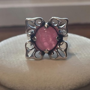 May include: A silver ring featuring a central round pink cabochon stone set within a square-shaped frame. The metal frame displays an openwork design with four symmetrical, curved, and pointed decorative elements at each corner.