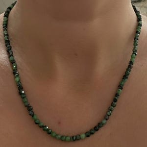 May include: A beaded necklace featuring small, faceted, round beads in varying shades of forest green, emerald, and dark charcoal gray. The beads are strung together in a continuous strand that rests around the neck, showcasing a reflective, polished surface.