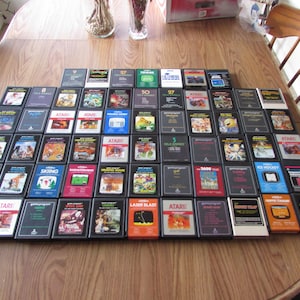 May include: A large collection of rectangular Atari video game cartridges arranged in a grid on a wooden table. Each cartridge features a black plastic casing with a colorful label on the front displaying various game titles and graphic illustrations.