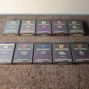 May include: A collection of ten rectangular black video game cartridges arranged in two rows. Each cartridge features the Tele-Games logo, a large number, and game titles including Asteroids, Breakaway IV, Capture, Gunslinger, Warlords, Auto Racing, Space Combat, Canyon Bomber, Speedway II, and Tank Plus.