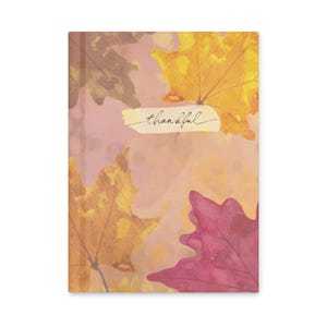 May include: A rectangular notebook with a soft pink background featuring watercolor-style autumn leaves in shades of yellow, orange, and deep magenta. A central cream-colored horizontal label displays the word thankful in black script.