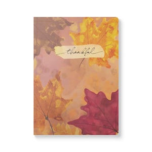 May include: A rectangular notebook featuring a watercolor-style autumn theme with scattered maple leaves in shades of yellow, orange, and deep red. A central cream-colored horizontal strip displays the word thankful in black cursive script.