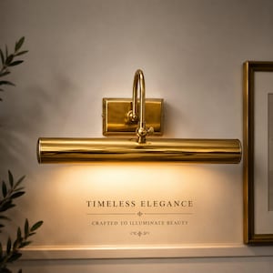 May include: A polished gold-tone metal picture light fixture with a rectangular wall mount and a cylindrical lamp head. The light emits a warm glow downwards. Below the fixture, the text reads: TIMELESS ELEGANCE CRAFTED TO ILLUMINATE BEAUTY.