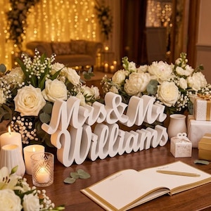 May include: A white, three-dimensional sign featuring the text Mrs & Mr Williams in a cursive font, displayed on a wooden table alongside white floral arrangements, lit candles, a gold pen, an open guestbook, and small gift boxes.