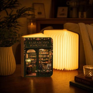 Op de afbeelding: Een boekvormige lamp met een miniatuurdiorama van een boekenwinkel genaamd Elizabeth's Bookstore, met een groene gevel, bloemaccenten en warme binnenverlichting, naast een open, accordeon-stijl verlichte boeklamp.