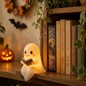 May include: A small, white, glowing ghost figurine sitting on a shelf edge, holding a tiny open book. The ghost has a smooth, rounded shape with two black eyes and a simple smiling mouth, emitting a soft, warm light from within.